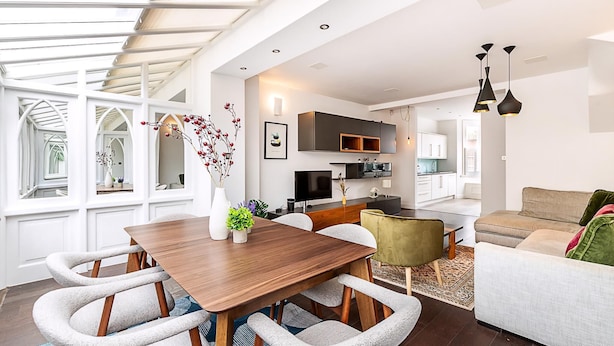 A modern living room with a wooden dining table, grey upholstered chairs, a sofa, and a kitchen in the background. - Elegant Chelsea Riverside Garden Retreat (London)