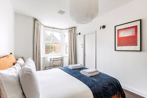 Ein Hotelzimmer mit Bett, Sessel, Schreibtisch, Fenster mit Vorhängen, Tür und einem gerahmten Bild an der Wand.