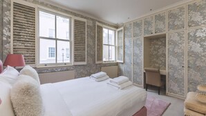 A bedroom with a large bed, a window, a radiator, a chair, and a wardrobe with floral wallpaper. - Stylish Chelsea Escape With Charm (London)