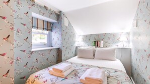 Un dormitori amb paper pintat floral, un llit amb almoadines, una finestra i una taula de nit amb llibres.
