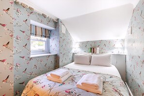 Ein Schlafzimmer mit floralem Wandpapier, einem Bett mit Kissen, einem Fenster und einem Nachttisch mit Büchern.