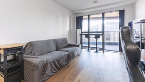 A modern living room with a sofa, a small table, and a chair. There is a large window with a view of buildings outside. - Large Bright Canal 1-bed With Balcony (London)