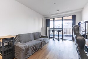 A modern living room with a sofa, a small table, and a chair. There is a large window with a view of buildings outside. - Large Bright Canal 1-bed With Balcony (London)