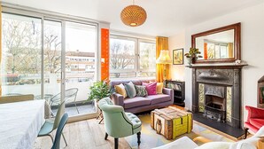 A living room with a sofa, a coffee table, a fireplace, and a large window with a view of buildings and trees. - Bright Bold Hackney Escape (London)