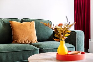 Ein grünes Sofa, ein braunes Kissen, eine gelbe Vase mit Blumen und ein roter Tablett mit einem weißen Gegenstand auf einem runden Tisch.