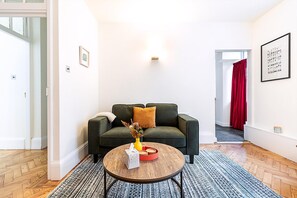 A living room with a dark green sofa, a round coffee table with a vase and a tissue box, a red curtain, and a framed picture on the wall. - Modern London Bridge Retreat (London)
