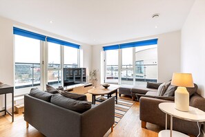 Ein modernes Wohnzimmer mit einer Couch, einem Sessel und einem kleinen Tisch mit einer Lampe. Große Fenster mit blauen Jalousien bieten Blick auf die Stadt.