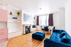 A modern living room with a fireplace, a blue sofa, a dining area, and a pink cabinet. - Colourful Three Bed Flat With Private Patio (London)