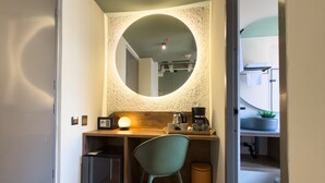 A compact room with a desk, chair, coffee maker, and a trash bin.