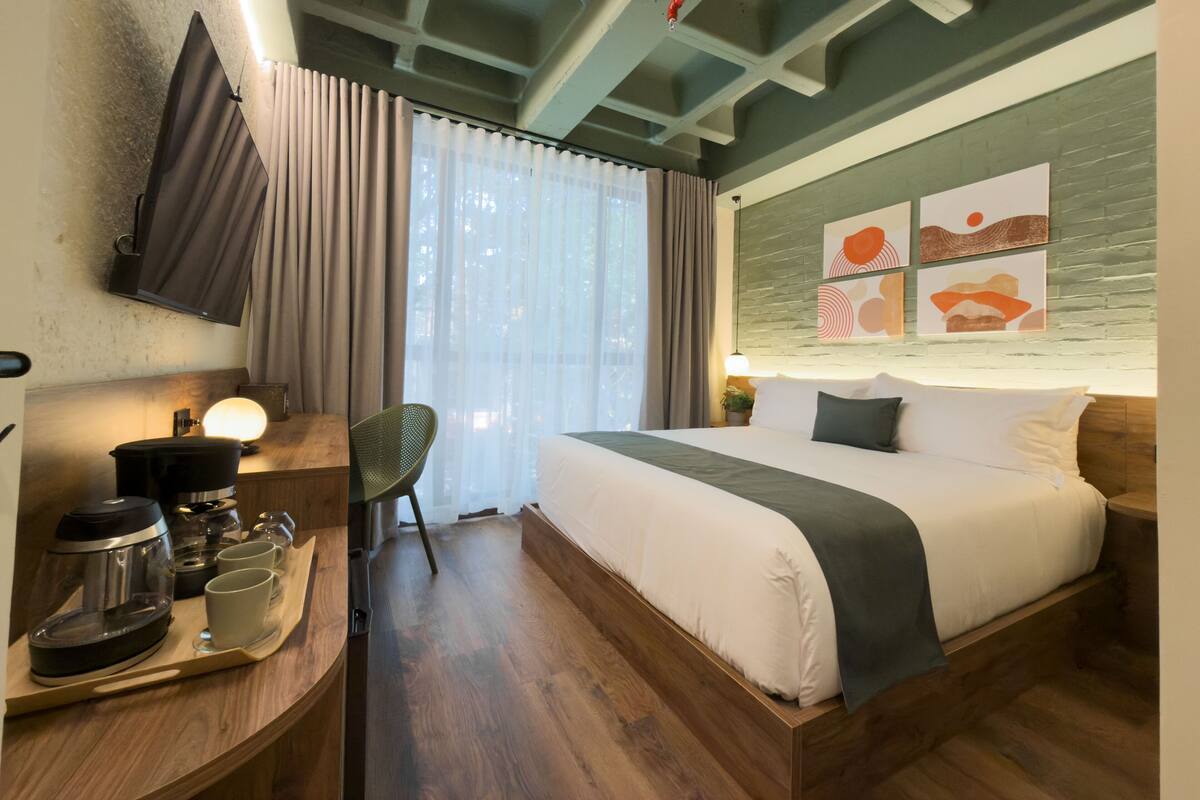 Una habitación de hotel con una cama, un escritorio con cafetera, una silla y obras de arte en la pared.