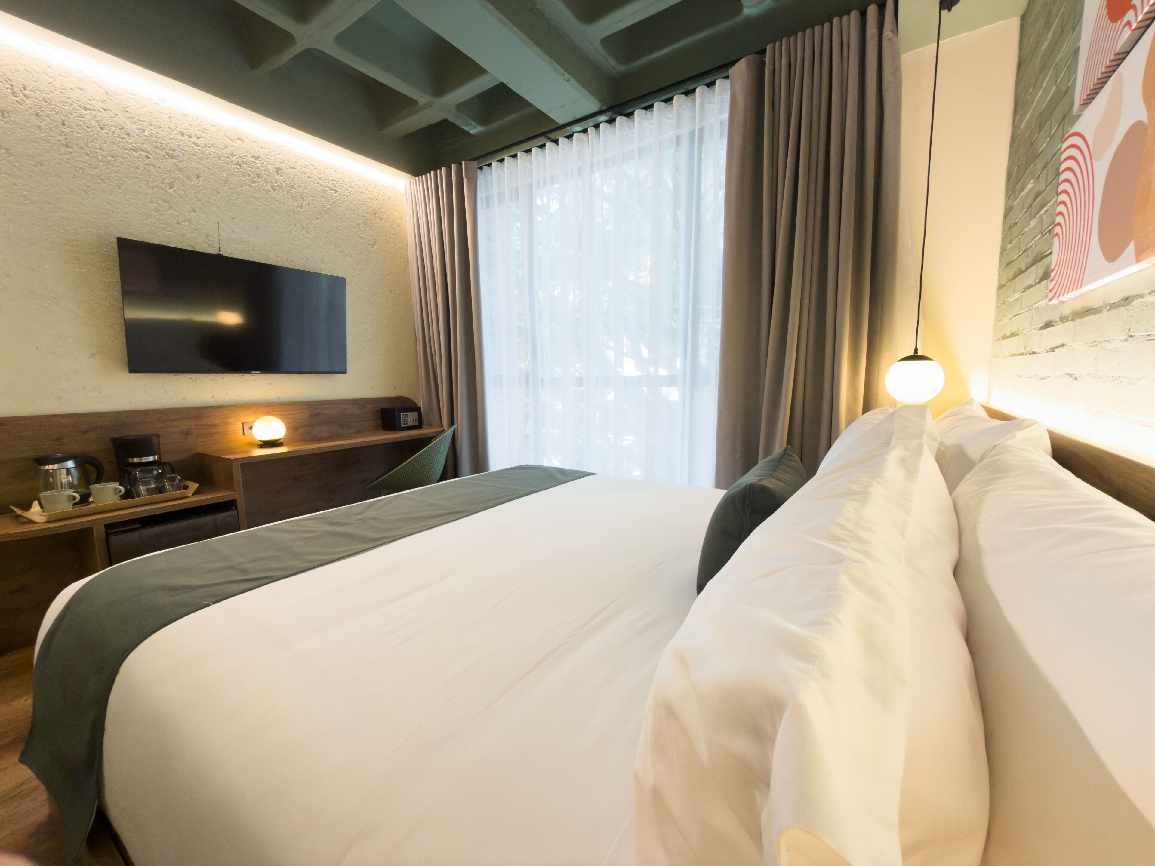 Una habitación de hotel con una cama, un televisor de pantalla plana montado en la pared, una cafetera y una ventana con cortinas.