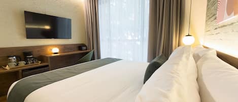 Una habitación de hotel con una cama, un televisor de pantalla plana montado en la pared, una cafetera y una ventana con cortinas.