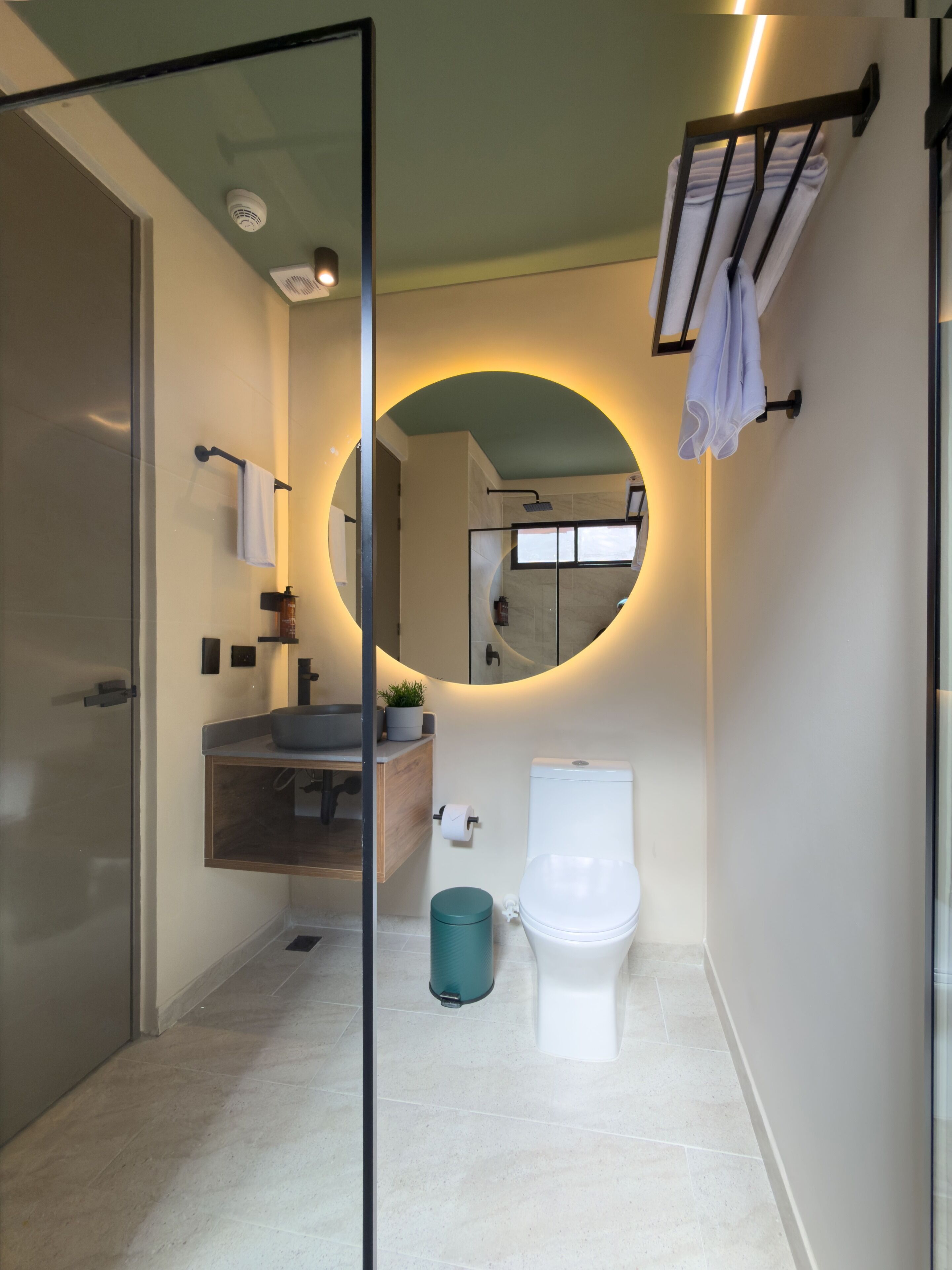 A modern bathroom with a glass shower enclosure, a round mirror with a circular light, a toilet, and a sink with a countertop.