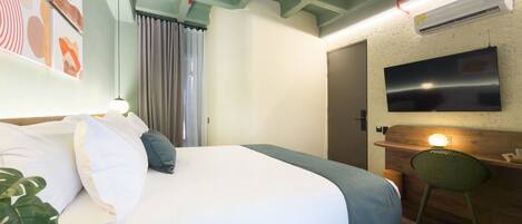 Ein modernes Hotelzimmer mit einem Bett, einem Schreibtisch mit Stuhl, einem an der Wand befestigten Fernseher und einer Pflanze.