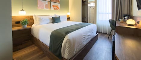 Habitación de hotel con cama, mesita de noche, lámpara, planta en maceta, una obra de arte en la pared y un escritorio con silla.