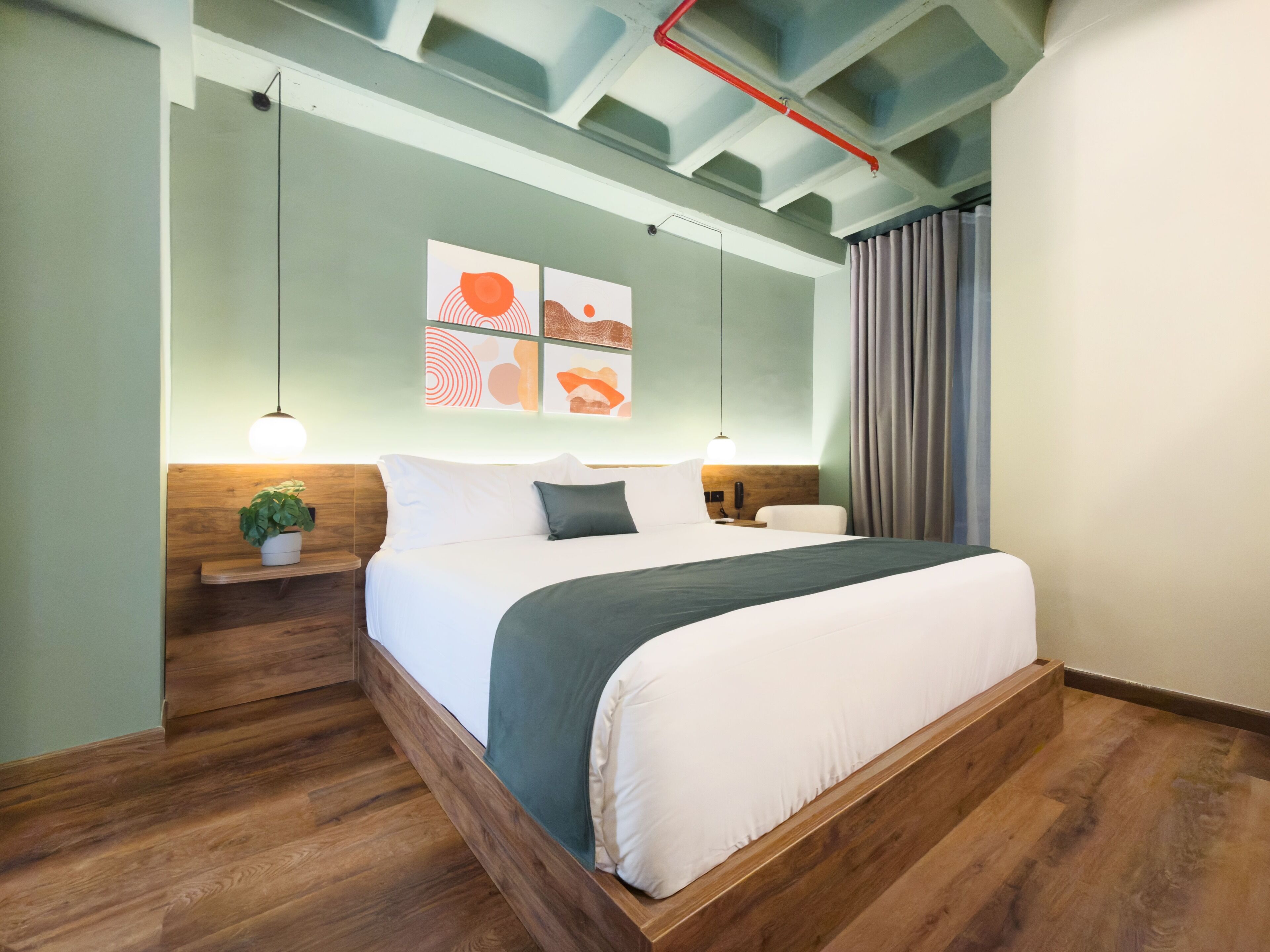 A modern hotel room with a wooden bed frame, a nightstand, a potted plant, and a ceiling with exposed beams.