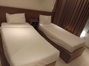 Deux lits simples avec du linge de lit blanc, une tête de lit en bois, une table de chevet et une fenêtre avec des rideaux.