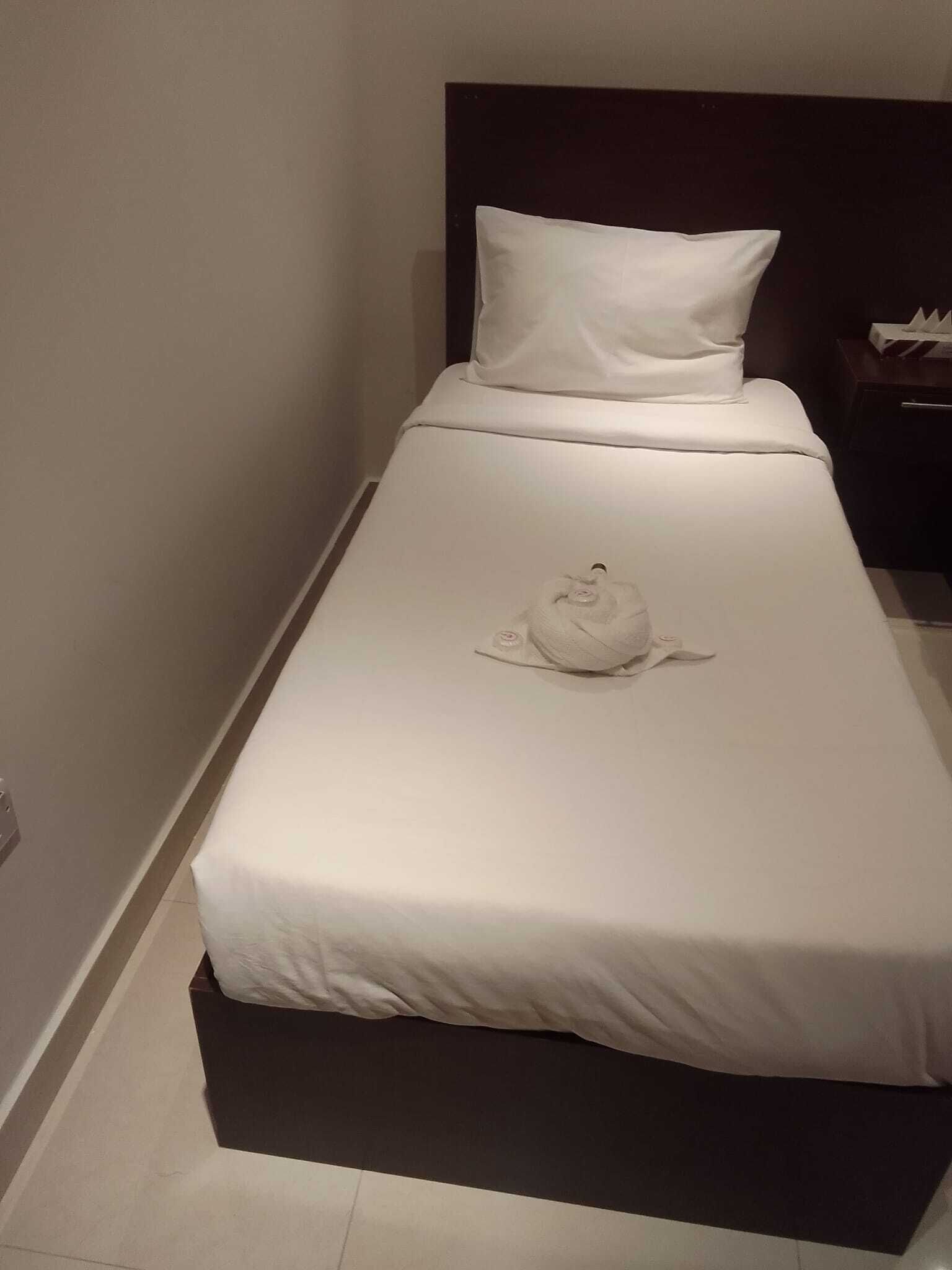 Una cama individual con colchón y almohada blancos, cabecera oscura y mesita de noche con lámpara.