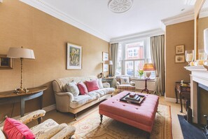 Un salon comprenant un canapé, un pouf, des fauteuils, une cheminée et une fenêtre avec des rideaux.