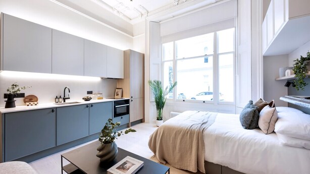 Een moderne slaapkamer met een groot raam, een bed met veel kussens, een nachtkastje met een boek, en een keukenhoek met kastjes en een aanrecht.