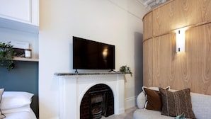Een moderne woonkamer met een flatscreen televisie boven een open haard, een bank met kussens en een kleine plant op de schoorsteenmantel.
