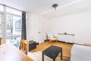 Ein Zimmer mit Holzboden, einem weißen Sessel, einem schwarzen Tisch, einem Bett, einem Heizkörper und einem Fenster mit Vorhang.