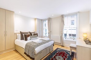 A bedroom with a bed, bedside table, lamp, window with curtains, and a view of buildings. - Elegant Pimlico Townhouse (London)