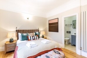 Une chambre à coucher comprenant un lit, des tables de chevet, une commode, une salle de bain avec des toilettes et un lavabo, et une porte donnant sur une autre pièce.