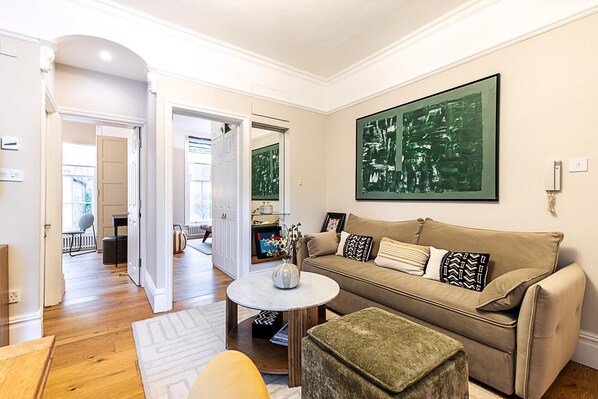 Un salon avec un canapé beige, une table basse ronde et un pouf vert. Un grand tableau abstrait orne le mur.