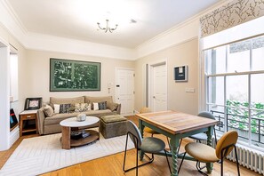 A living room with a sofa, coffee table, and a dining table with chairs. There is a large window with a view of greenery outside. - Stylish Flat Near Denmark Hill (London)
