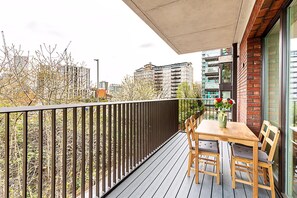 A balcony with a wooden table and chairs, a potted plant, and a view of buildings and trees. - Stratford Flat Near Abba Arena (London)