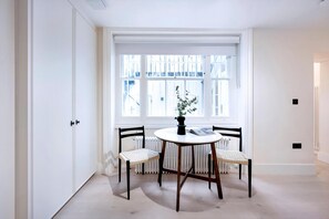 Een kamer met een witte radiator, een raam met witte kaders, een ronde tafel met een zwarte vaas en twee rieten stoelen.