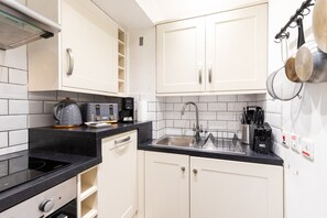 A kitchen with white cabinets, a black countertop, a stainless steel sink, a coffee maker, and a kettle. - Snug Holland Park Stay (London)