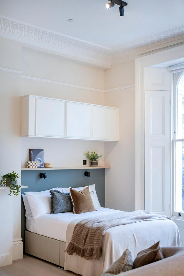 Ein Schlafzimmer mit einem Bett, weißen Schränken darüber, einem Fenster und einer Pflanze an der Wand.