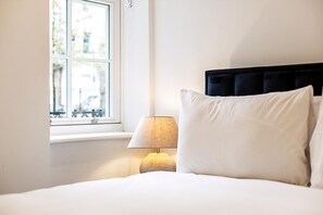 Ein Schlafzimmer mit einem Bett, weißen Kissen, einem Nachttisch mit Lampe und einem Fenster mit Blick auf Gebäude.