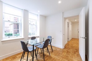 A modern room with a glass table and chairs, large windows, wooden flooring, and a hallway leading to another room. - Modern and Elegant Apartment in West Kensington (London)