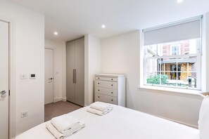 A bedroom with a bed, a dresser, a window with a view of buildings, and a door leading to another room. - Modern and Elegant Apartment in West Kensington (London)