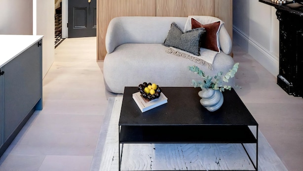 A modern living room with a sofa, a coffee table with a book and fruit, a decorative ceiling, and a wooden panel wall. - Normandy Court 11 (London)