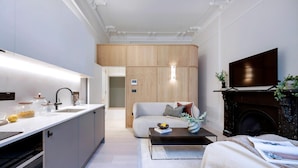 A modern kitchen with a built-in oven, a dining area with a sofa and coffee table, and a living room with a fireplace and TV. - Normandy Court 11 (London)