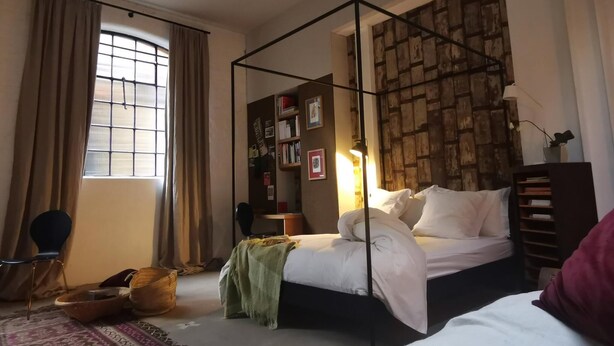 A bedroom with a four-poster bed, a bookshelf, a window with curtains, and a patterned rug. - Architect Designed Studio Borough (London)