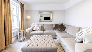 A living room with a sectional sofa, a tufted ottoman, a side table, a floor lamp, and a mirror on the wall. - Stylish Marylebone City Apartment (London)