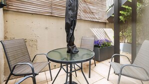 A patio area with a round glass table, two metal chairs, a black umbrella, and a planter with purple flowers. - Chelsea Retreat With Private Patio (London)