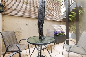 Eine Terrasse mit einem runden Glastisch, zwei Metallstühlen, einem schwarzen Sonnenschirm und einem Pflanzgefäß mit violetten Blumen.