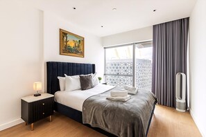 A bedroom with a bed, a nightstand, a heater, a window with curtains, and a painting on the wall. - Stylish 2-bed by The O2 With Balcony (London)