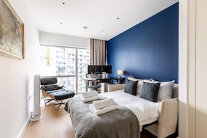 A hotel room with a bed, a desk, a chair, a TV, and a large window with a view of buildings. - Stylish 2-bed by The O2 With Balcony (London)
