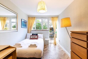 Ein Schlafzimmer mit Bett, Nachttisch, Lampe, Kommode, einem Fenster mit Vorhängen und Blick ins Freie.