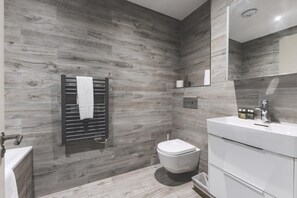 A modern bathroom with a wood-textured wall, a white toilet, a black towel rack, and a white vanity with a mirror. - Stylish Wimbledon Flat With Balcony (London)