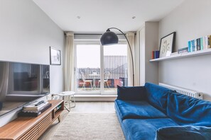A modern living room with a blue sofa, a TV, a wooden coffee table, and a large window with a view of a balcony. - Stylish Wimbledon Flat With Balcony (London)