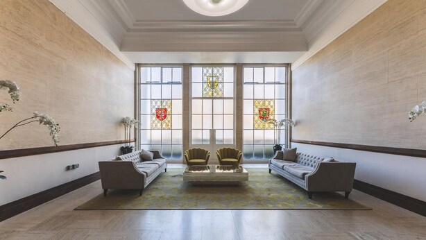 Un hall d’entrée spacieux avec un plafond haut, de grandes fenêtres ornées de vitraux, deux canapés et une table basse.