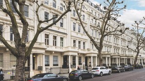 A row of traditional buildings with uniform architecture, a clear sky, and parked cars along the street. - Elegant South Kensington Retreat (London)